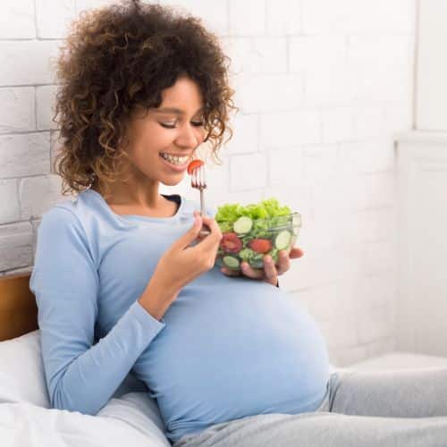 Black pregnant woman eating salad in bed